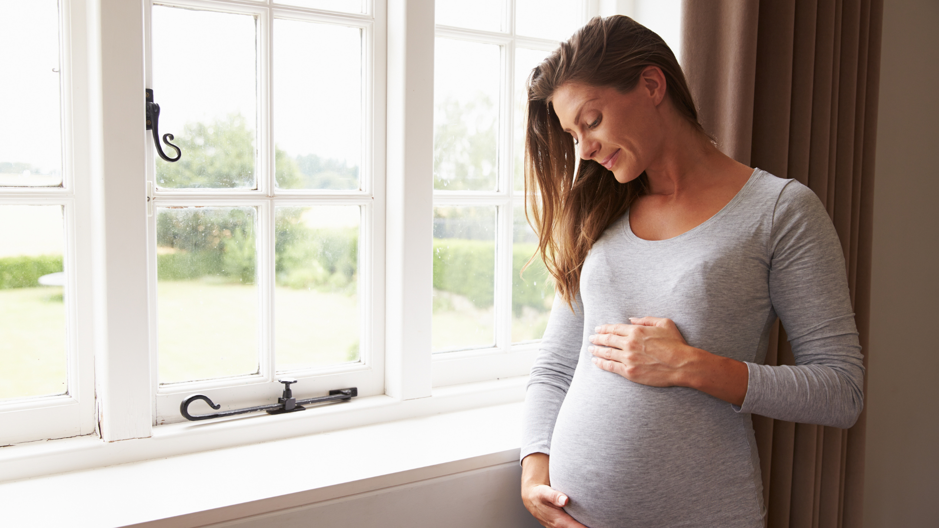 Pregnant woman holding her belly while looking outside the bedroom window