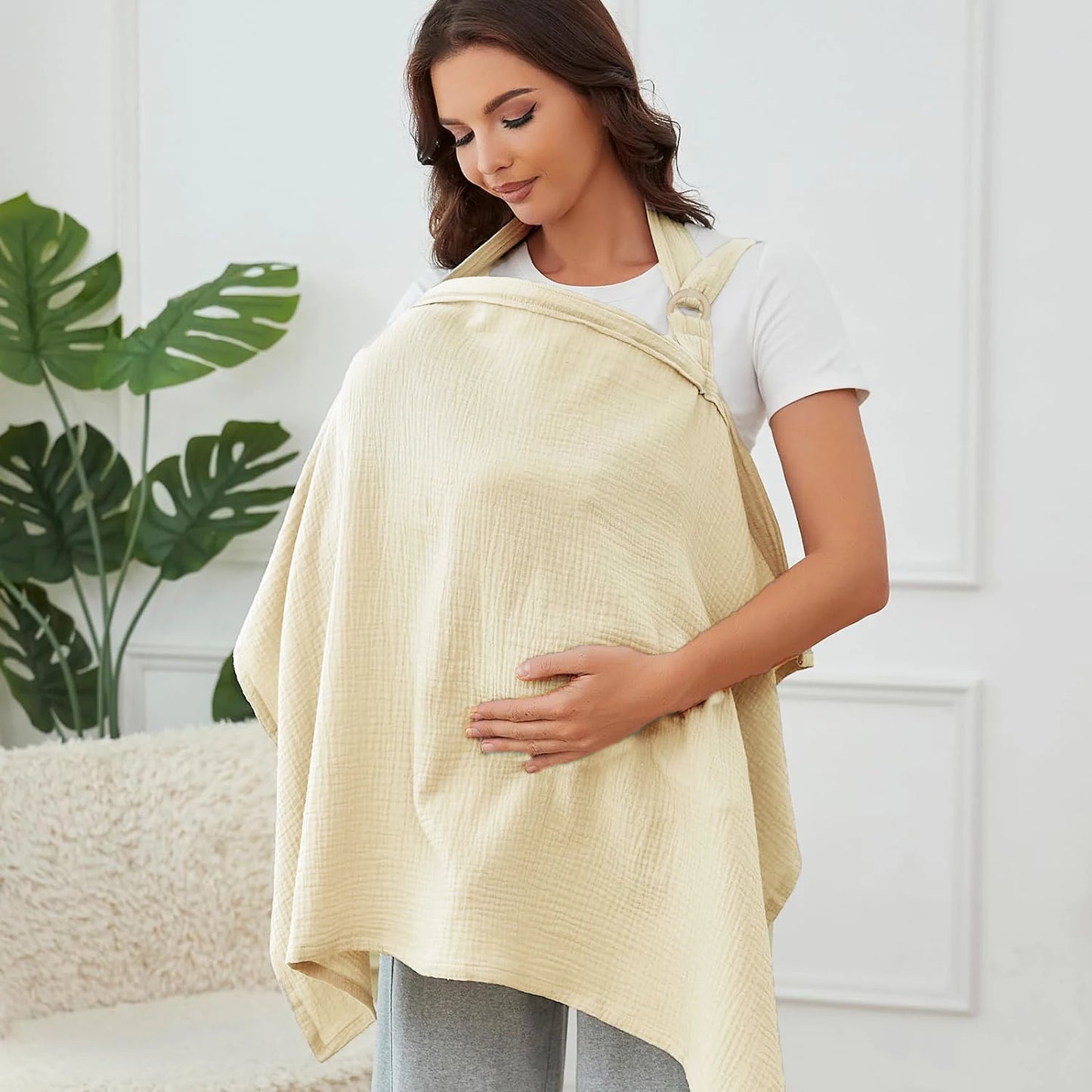 Woman wearing a beige baby carrier in a home setting with a plant in the background