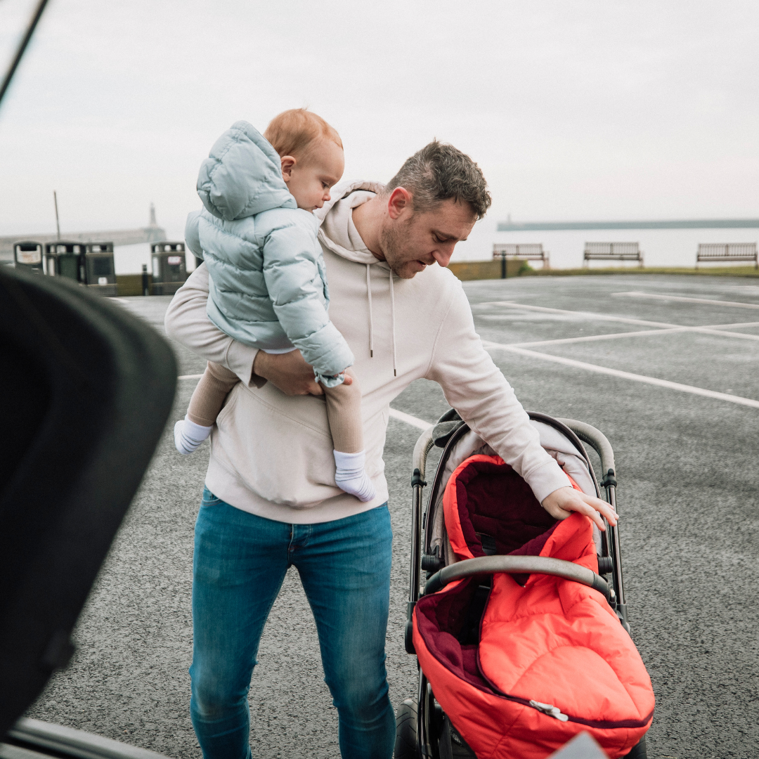 Man holding a child and pushing a red stroller in a parking lot.