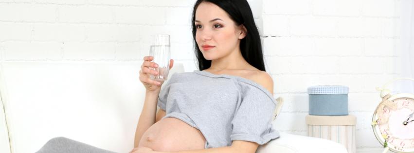 A pregnant woman sitting on a couch, looking comfortable and relaxed, with a glass of water and healthy snacks nearby.