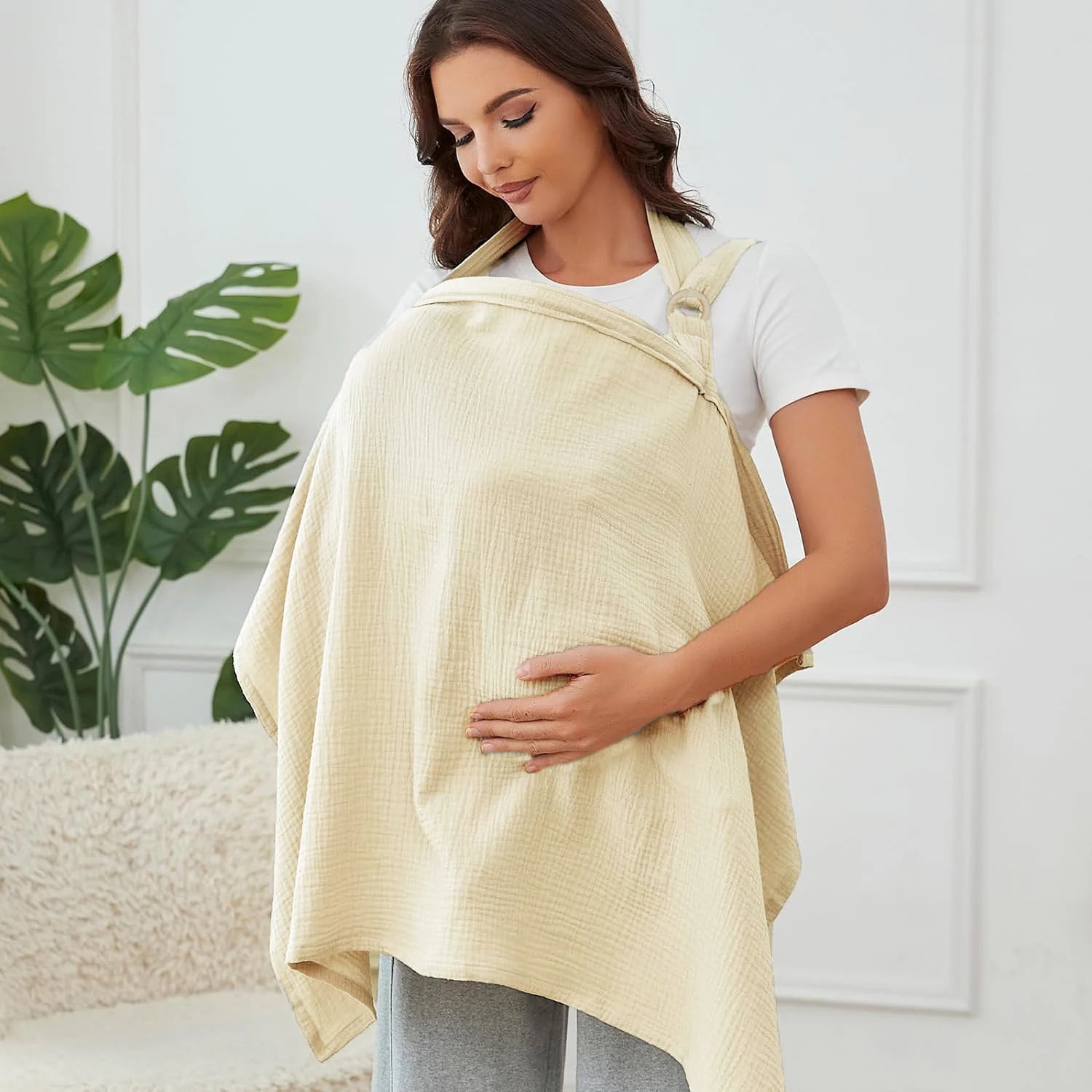 Woman wearing a beige baby carrier in a home setting with a plant in the background