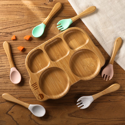 Wooden children's plate with divided sections on a wooden surface, surrounded by colorful spoons and forks.