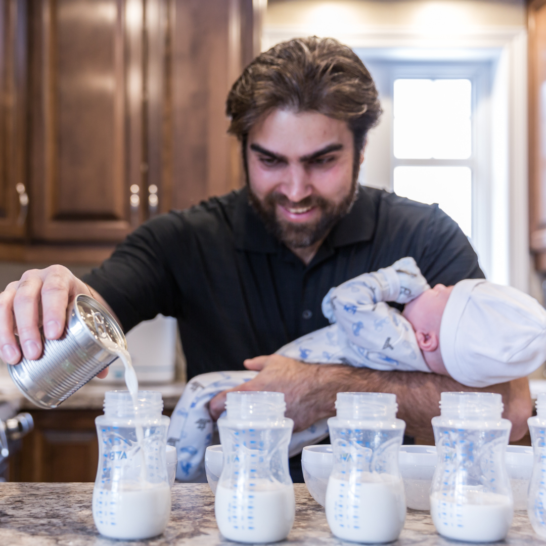 Man pouring formula into baby bottles while holding a baby in a kitchen.