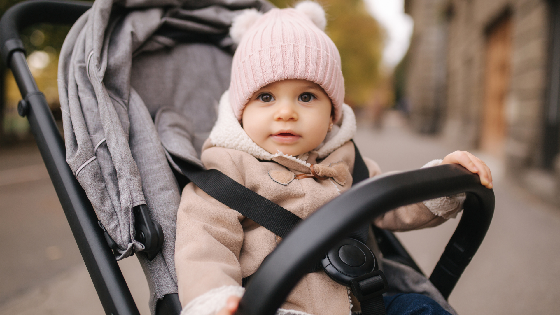 Baby in a stroller wearing a pink hat and coat on a street.