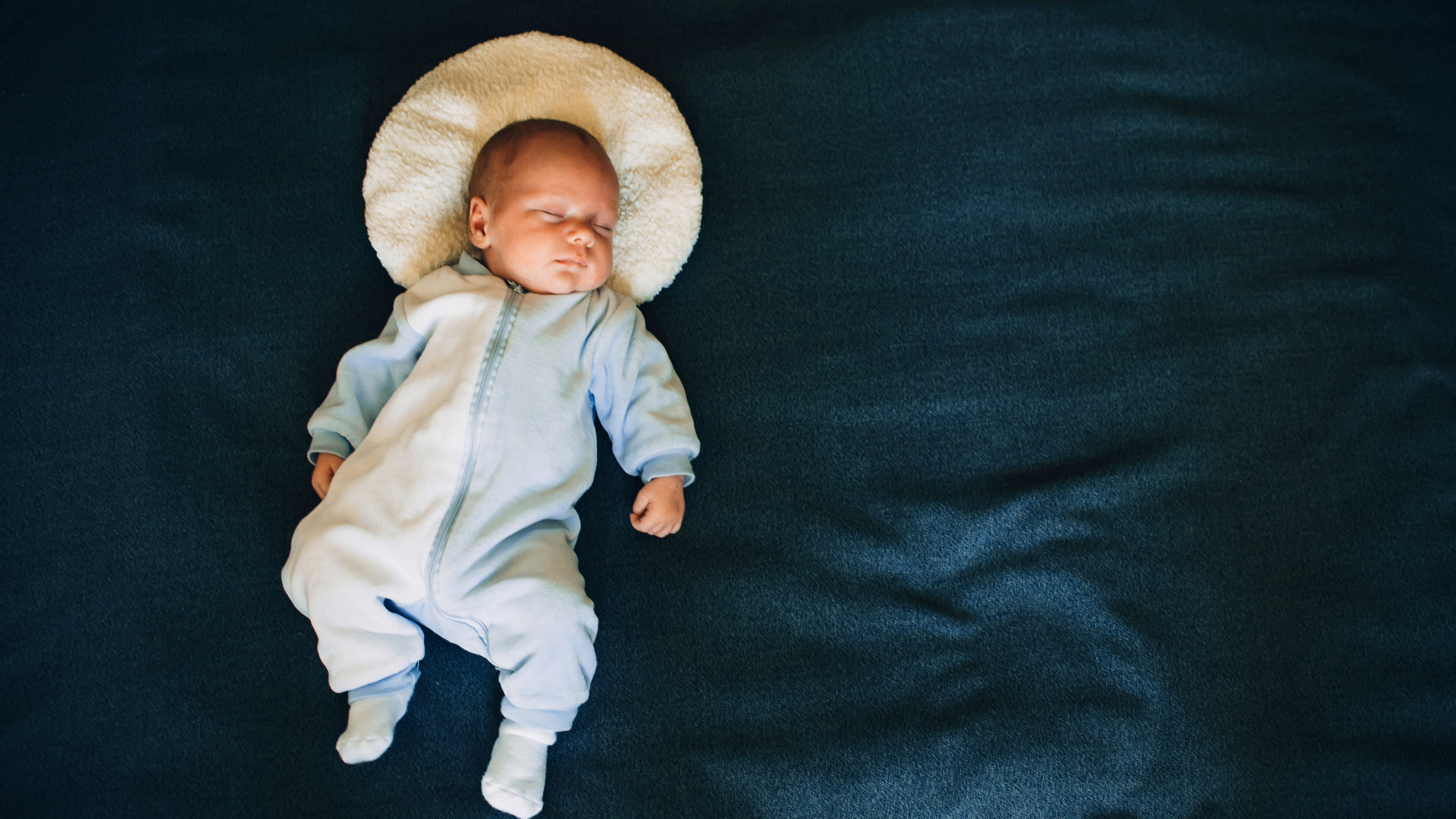 Baby in a light blue onesie with a white hat on a dark blue blanket